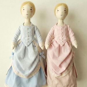 Op de afbeelding: Twee handgemaakte stoffen poppen, elk met blond haar en een eenvoudig gezicht. De ene pop draagt een blauw-wit gestreepte jurk met kanten rand, de andere een roze-wit gestreepte jurk. Beide jurken hebben een vergelijkbaar ontwerp.