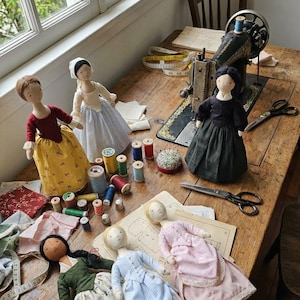 May include: Handmade fabric dolls dressed in historical clothing are arranged on a wooden table with sewing supplies. A vintage sewing machine, spools of thread, fabric scraps, and patterns complete the scene. The dolls' outfits include a red bodice and yellow skirt, and a black dress.