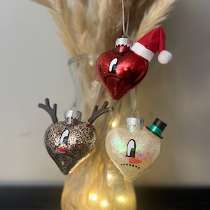 May include: Three heart-shaped Christmas ornaments with painted faces. One ornament is red with a Santa hat, one is brown with glitter and reindeer antlers, and one is white with glitter and a snowman hat.