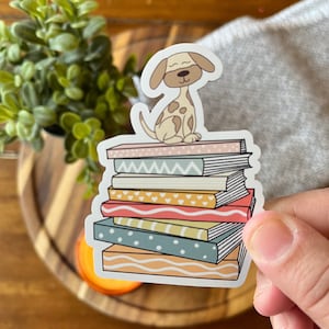 Pode incluir: Um adesivo de um cão castanho e branco sentado em cima de uma pilha de livros. Os livros têm padrões diferentes, incluindo corações, listras e ondas. O adesivo está sobre uma superfície de madeira.