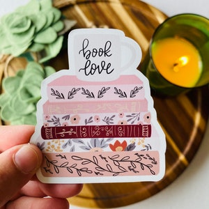 以下が含まれることがあります： 本の積み重ねに花柄のデザインと「book love」と書かれたコーヒーカップが描かれたステッカーです。