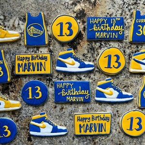 May include: A collection of 13th birthday cookies decorated with basketball themes. The cookies feature a basketball jersey, basketball shoes, and the number 13. The cookies are decorated in blue, yellow, and white.