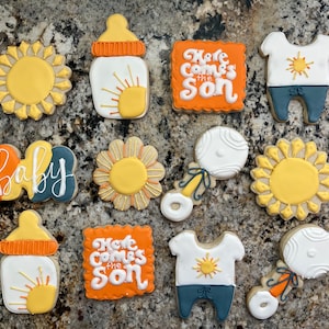 Peut inclure: Un ensemble de 10 biscuits de fête prénatale décorés sur le thème du soleil. Les biscuits représentent un biberon, un hochet, une grenouillère, un soleil et le texte "Here comes the son".