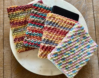 May include: Four colorful, hand-crocheted washcloths in various shades of red, yellow, blue, and green, arranged on a white marble plate. The washcloths have a textured appearance and are displayed on a wooden surface.