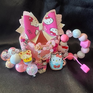 May include: A pink glittery container holding a Hello Kitty hair bow, a beaded bracelet with pink and white beads, and various charms including a Hello Kitty cupcake, a Hello Kitty in a blue mug, and a charm with the text be kind.