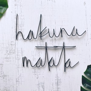 May include: Black wire wall art with the words "Hakuna Matata" on a white wood background with green leaves on either side.