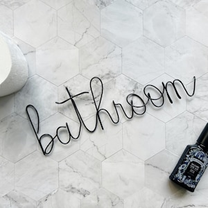 May include: A roll of white toilet paper, a black wire word "bathroom", and a dark bottle with a floral design are arranged on a white and grey hexagonal tile surface. The items suggest a bathroom theme.