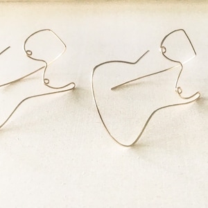 May include: A pair of gold-toned earrings shaped like a guitar. The earrings are made of thin wire and have a simple, minimalist design.