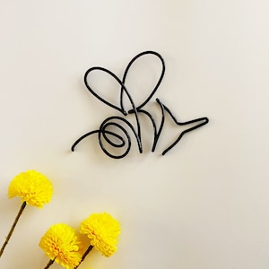 May include: A black wire sculpture of a bee on a white background with yellow flowers in the bottom left corner.