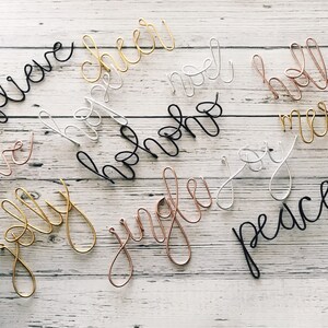 May include: Assortment of wire word art in various colors, including black, gold, silver, and copper. Words like "believe", "cheer", "noel", "holly", "merry", "love", "hohoho", "joy", "jingle", and "peace" are displayed.