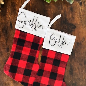 Puede incluir: Dos calcetines de Navidad de cuadros rojos y negros con tapas de tela blanca. Los calcetines están personalizados con los nombres "Griffin" y "Bella" en cursiva negra.