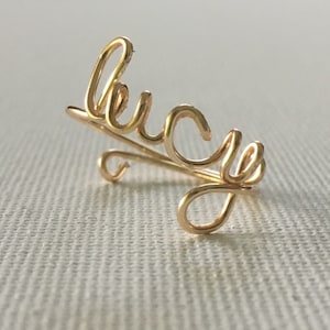 Puede incluir: Anillo de alambre dorado con la palabra "Lucy" escrita en cursiva.