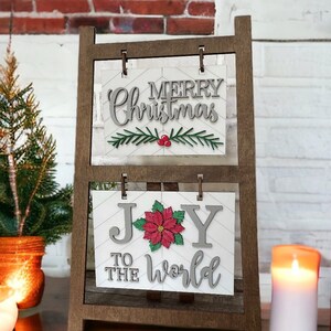 May include: A wooden easel with two signs. The top sign says "Merry Christmas" in gray lettering with a green sprig and red berries. The bottom sign says "Joy to the World" in gray lettering with a red poinsettia.