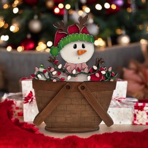 May include: A wooden Christmas decoration featuring a snowman wearing a red hat with antlers and a green beanie. The snowman is in a brown basket decorated with string lights. The background includes a Christmas tree and wrapped gifts.