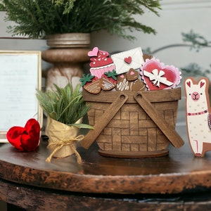 May include: A wooden picnic basket filled with Valentine's Day themed decorations, including a cupcake, hearts, and a llama. The basket is sitting on a wooden table.