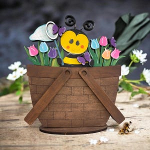 May include: A wooden decorative basket with a bee and butterfly theme. The basket is filled with colorful tulips and butterflies. The bee is yellow and black, holding a butterfly net. The basket is brown with wooden handles.