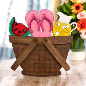 May include: A wooden picnic basket with decorative cutouts of a watermelon slice, pink polka-dot flip-flops, and a yellow lemonade pitcher. The basket has a woven design and wooden handles, evoking a summer theme.