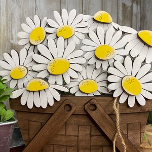May include: A wooden basket filled with white daisy flowers with yellow centers. The daisies are arranged in a bouquet and are sitting in a brown wooden basket with a woven pattern.