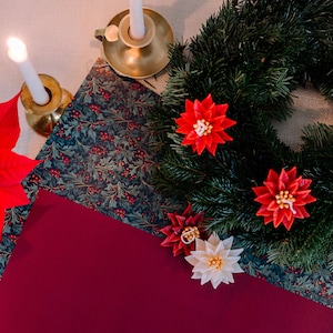 Könnte beinhalten: Ein Weihnachtskranz mit roten Poinsettien-Dekorationen. Zwei brennende Kerzen in Messinghaltern stehen auf einer gemusterten Oberfläche. Eine tiefrote Oberfläche und gemustertes Papier mit Beeren sind ebenfalls sichtbar.