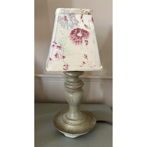 May include: A table lamp with a tapered, rectangular fabric lampshade featuring a floral pattern in shades of pink, green, and cream. The lamp base is made of a light green, distressed-finish material with a turned, pedestal-style design.