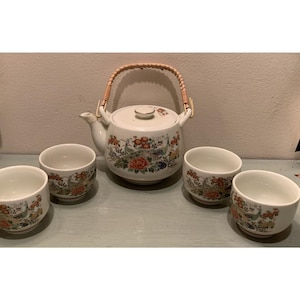 May include: A white ceramic tea set with a teapot and four matching cups. The teapot has a woven handle and a floral and bird design in red, orange, green, and blue. The cups have a similar design.