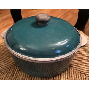 May include: A round teal cooking pot with a matching lid and a central gray knob. The pot features a metallic silver rim and two side handles, showcasing a smooth, enameled finish on the exterior surface of the lid and the main body.