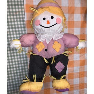 May include: A pink and yellow plush scarecrow doll with a white face, black eyes, and an orange nose. The scarecrow is wearing a pink shirt with yellow patches, black trousers, and yellow shoes.