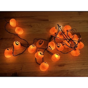 May include: A string of orange pumpkin-shaped lights connected by a black wire. Each plastic pumpkin features a carved jack-o'-lantern face with black detailing. The lights are illuminated, casting a warm glow from within each individual pumpkin.