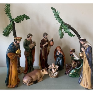 May include: A ceramic nativity scene featuring figures of the Holy Family, the Three Wise Men, and a cow. The figures are dressed in robes of blue, gold, green, and brown. Palm tree decorations are also included.