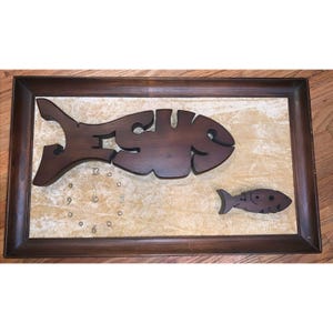 May include: A wooden wall clock shaped like a fish with the word "Jesus" carved into it. The clock is framed in a dark wood frame and has a beige background.
