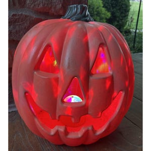 May include: A molded orange plastic jack-o'-lantern with a dark gray stem. The pumpkin features cutouts for triangular eyes, a triangular nose, and a wide, jagged mouth. It is illuminated from within by a rotating light displaying red and blue hues.