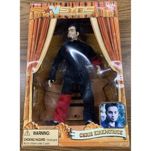 May include: An *NSYNC Chris Kirkpatrick collectible marionette doll in its original packaging. The doll is dressed in a black and red jumpsuit. The box features the *NSYNC logo and the text "Collectible Marionette". The box also includes a warning about small parts.