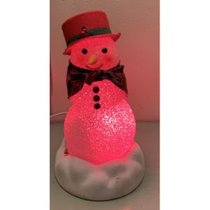 May include: A light-up snowman figurine with a red top hat, a plaid scarf, and three black buttons. The snowman has a carrot nose and is illuminated with a red glow. It sits on a white, cloud-shaped base.