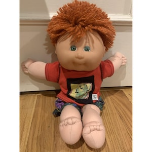 May include: A vintage Cabbage Patch Kid doll with a red shirt, colorful shorts, and orange yarn hair. The doll has green eyes and is sitting on a wooden floor. The shirt has a small patch with a design and the letters "cpk".