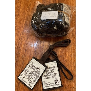 May include: A package of 10 brown cords and two scapulars. The scapulars have printed text and illustrations, including the words "a sign of Salvation" and "Our Lady of Mt. Carmel". The text "soever dies clothed in this Scapular shall not suffer eternal fire" is also visible.