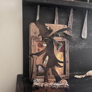 May include: A rustic, handcrafted metal figure of a man in a top hat and coat, holding a cane. The figure is mounted on a wooden base with a small wooden frame behind it. The background includes antique spoons.