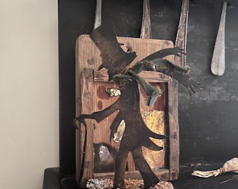 May include: A handcrafted metal figure of a man in a top hat and coat, holding a cane. The figure is mounted on a wooden base with a small wooden frame behind it. The background includes antique spoons.