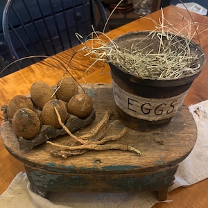 Puede incluir: Un taburete de madera rústico con una base pintada de azul, decorado con una colección de huevos marrones en una caja de cartón, ramitas secas y una pequeña olla marrón con un asa de alambre llena de hierba seca y la palabra "EGGS" escrita en el lateral.