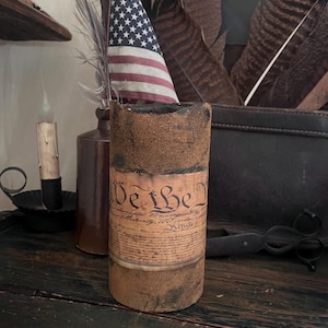 May include: A white ceramic mug with a black handle and the text "But First, Coffee" in a bold, black font. The mug is set against a blurred background, suggesting a cozy setting.