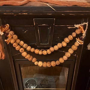 Puede incluir: Una repisa de chimenea negra está decorada con una guirnalda de cuentas de madera y rodajas de naranja secas. Una tela naranja drapeada y una tela beige están drapeadas sobre la parte superior de la repisa. La chimenea tiene una puerta de cristal.