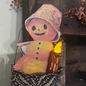 Puede incluir: Decoración de Halloween de estilo vintage con una figura de calabaza sonriente con sombrero de paja. La calabaza sostiene una vela en una cesta tejida con un patrón en blanco y negro. La vela tiene una luz amarilla parpadeante.