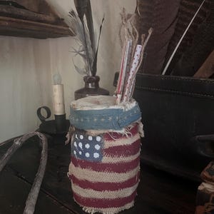 Puede incluir: Un tarro cilíndrico de arpillera decorado con un diseño de bandera estadounidense cosida. El tarro tiene una banda de tela azul con estrellas blancas alrededor de la parte superior. Se colocan plumas y papel enrollado dentro del tarro.