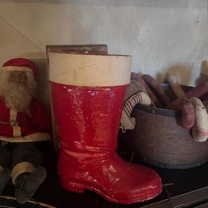 May include: A glossy red decorative boot with a cream-colored cuff. The boot is part of a holiday display, with a Santa Claus figure and a wooden container filled with fabric items visible in the background.