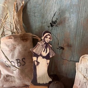 Op de afbeelding: Een houten figuur in vintage stijl van een vrouw in een lange jurk en een muts. De figuur staat op een houten voet en is omringd door andere decoratieve items, waaronder een jute zak met het woord "COBS" erop gedrukt, een grafsteen en een paar ronde, donkere objecten.