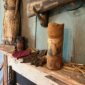 May include: A rustic, cylindrical candle with a textured, brown exterior and a vintage illustration. The candle sits on a wooden shelf with other decorative items, including a small owl figurine, dried leaves, and straw. A vintage rifle is mounted on the blue wall behind the shelf.