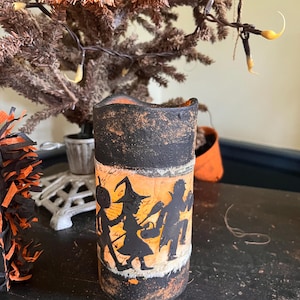 May include: A black and orange Halloween candle with a distressed finish. The candle features a silhouette of three trick-or-treaters, including a witch, a vampire, and a ghost.