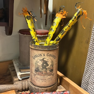 Op de afbeelding: Een cilindervormig metalen blik met het label Goblin's Goodies Old Fashioned Halloween Candy en een heksenillustratie, met drie gele stokjes met zwarte patronen en oranje franjes, plus twee rood-zwart gestreepte stokjes aan de voorzijde.