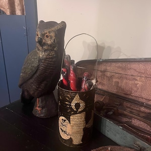 Op de afbeelding: Een decoratief uilbeeld staat naast een zwarte en gouden kandelaar gevuld met rode kaarsen. De kandelaar heeft een kaarsontwerp met tekst en een heksenillustratie. Een kist in vintage-stijl en een mand met decoratieve items zijn ook zichtbaar.