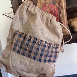 May include: A small, brown burlap drawstring bag with a plaid fabric pocket. The bag is hanging from a wooden surface.