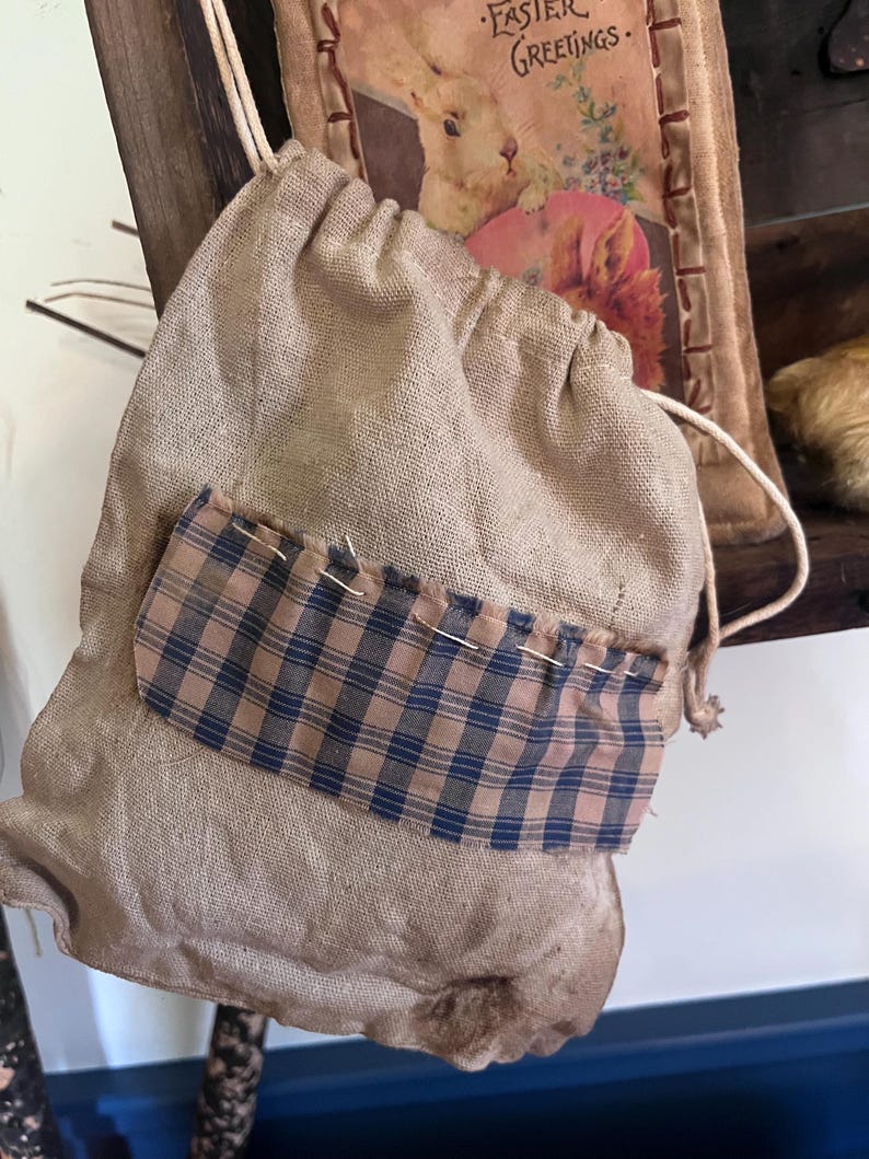 May include: A small, brown burlap drawstring bag with a plaid fabric pocket. The bag is hanging from a wooden surface.